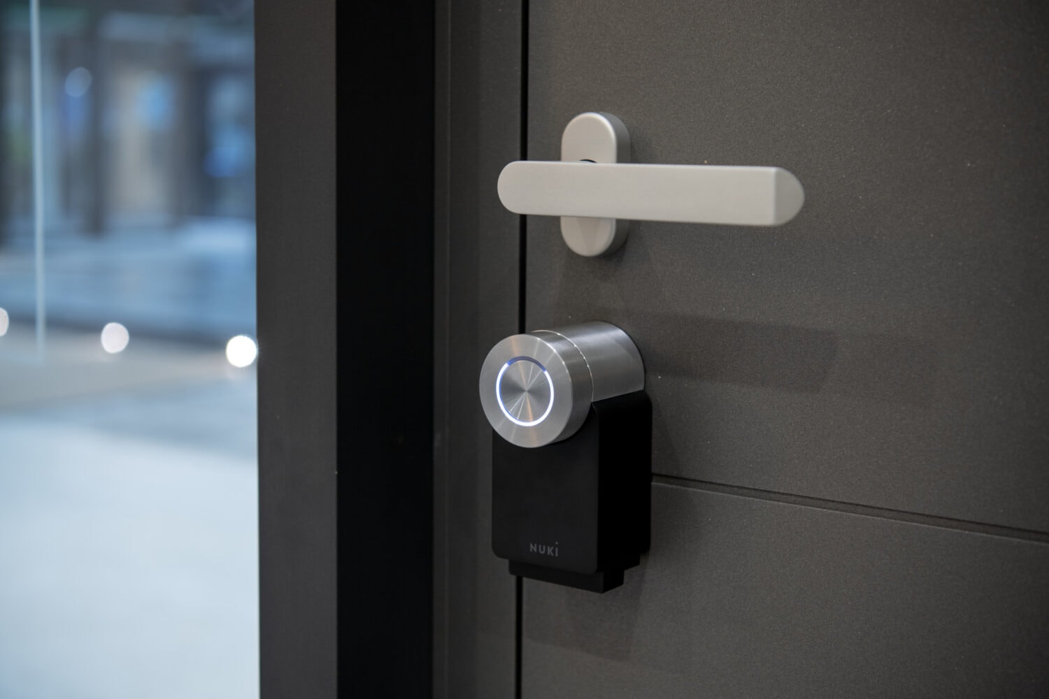 A modern smart lock is installed on a gray front door, featuring a sleek metallic handle and a circular illuminated control on the black smart lock device.