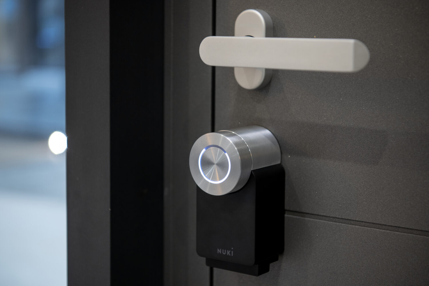 A close-up of a modern electronic smart lock with a silver knob and a black body labeled Nuki, installed below a sleek silver door handle on a dark gray front door.