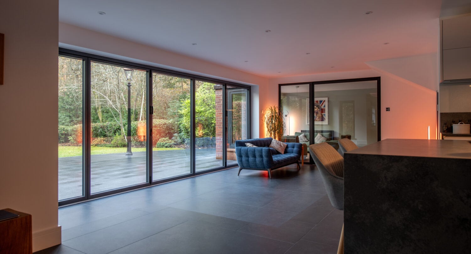 A modern living room with large sliding doors, crafted using bespoke glass solutions, opens to a garden and features contemporary furniture and warm indoor lighting.