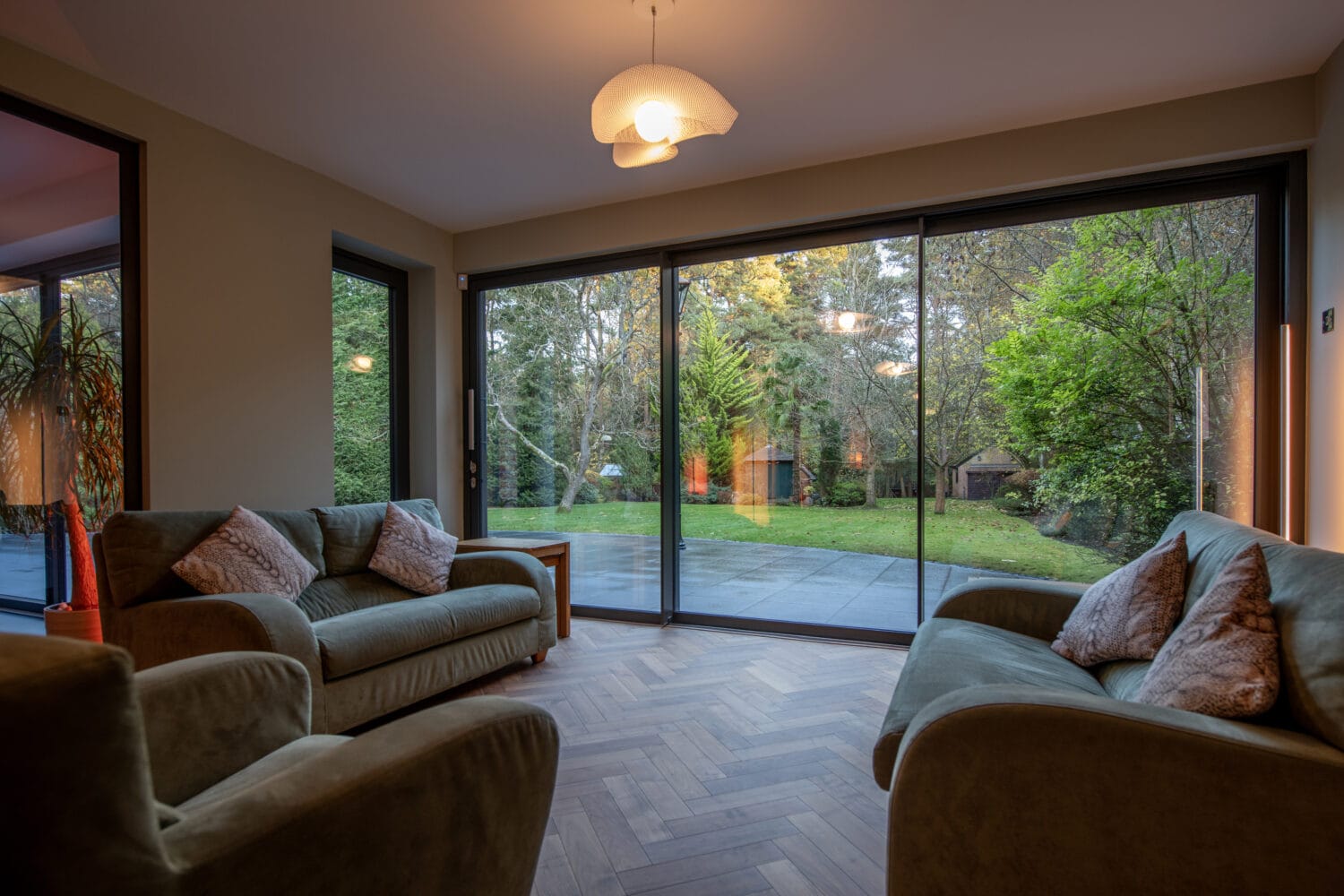 Spacious living room with two sofas, herringbone wood floor, and large sliding doors featuring bespoke glass solutions, offering a stunning view of the green garden with trees and outdoor lighting at dusk.