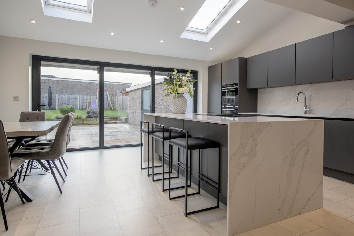 Modern kitchen with large marble island, black barstools, dark cabinets, and built-in appliances. Bespoke glass solutions like bifold doors and skylights fill the room with natural light and open to a patio and garden.