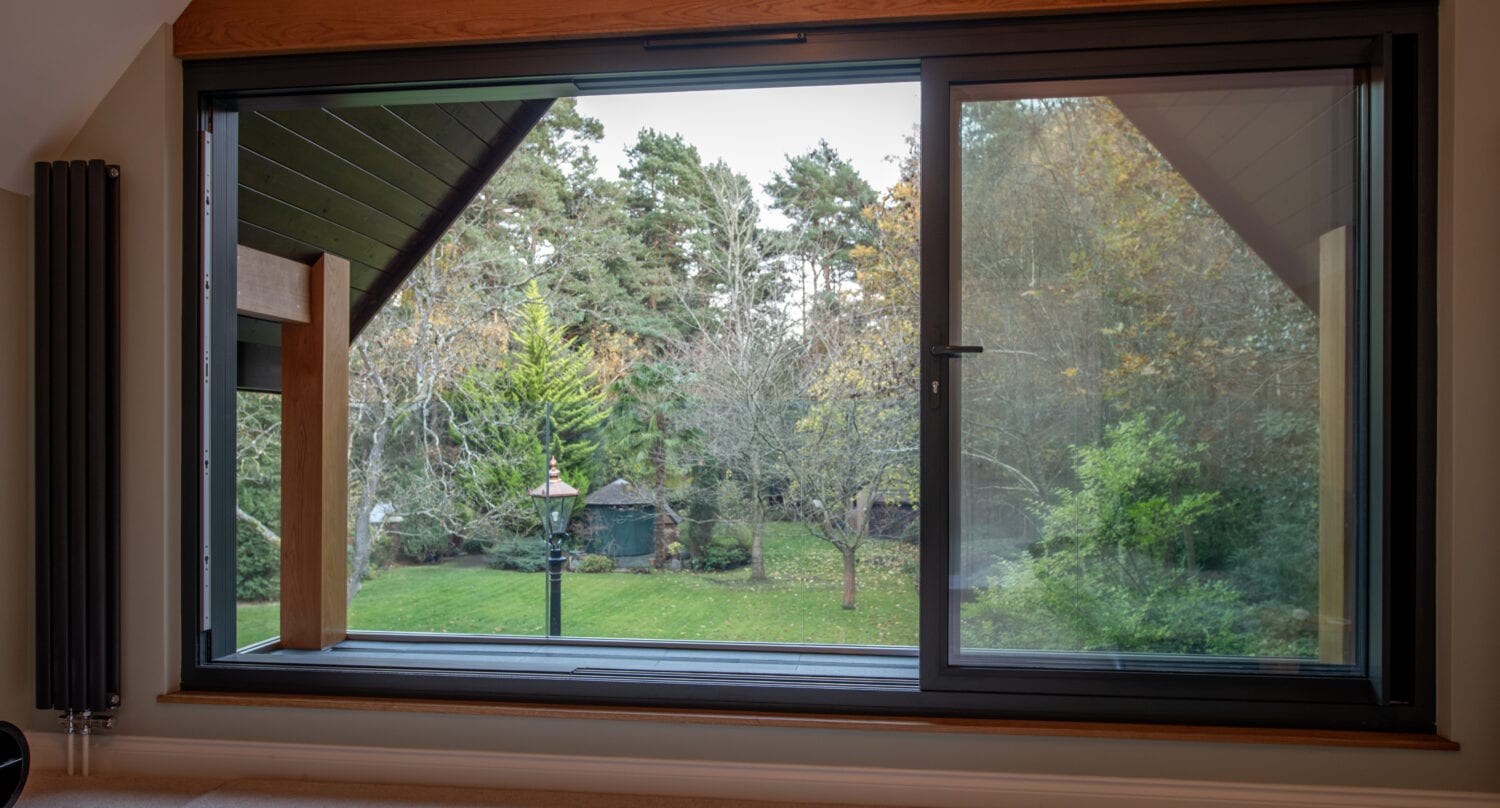 Large sliding glass doors in a modern room open to a lush green garden with trees, grass, a lamppost, and a gazebo. The view is beautifully framed by wooden beams and a slanted glass roofing outside.