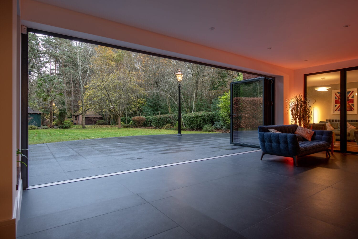 A modern living room with bespoke glass solutions features large folding glass doors that open to a spacious patio and green garden. A blue sofa sits near the doorway, and warm indoor lighting contrasts with the outdoor view.