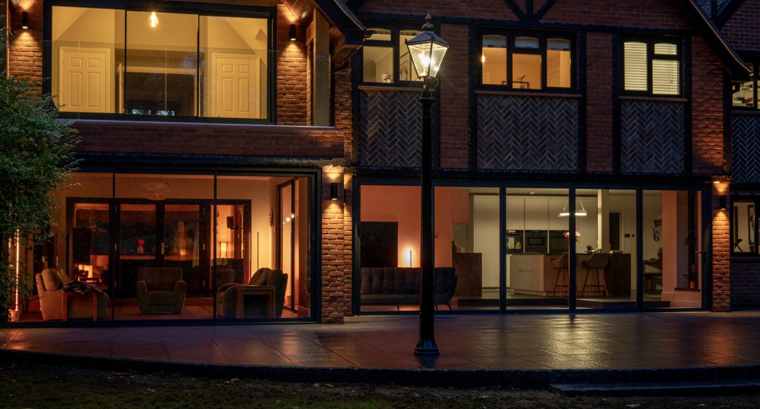 A modern house with large glass windows and bifold doors is warmly lit from inside at dusk. The patio and garden are visible in the foreground, with a vintage-style streetlamp near the entrance.