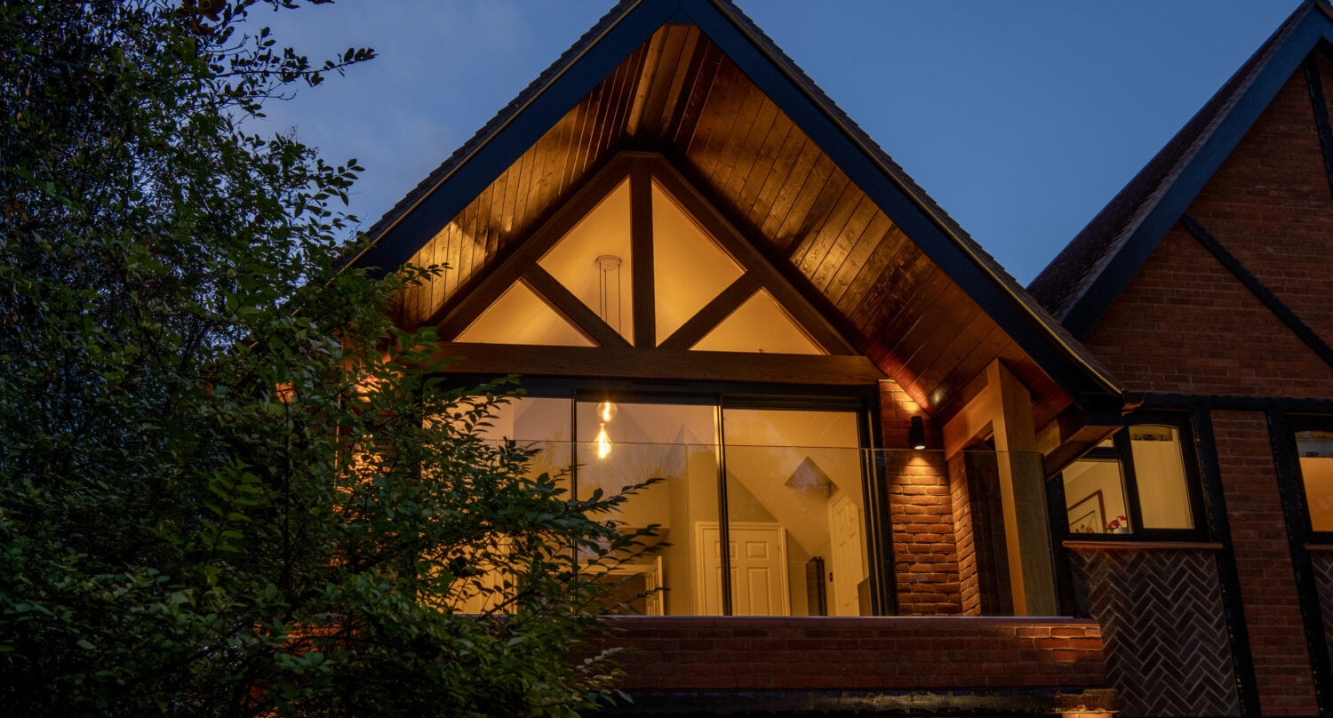 A modern house with large triangular windows, wooden beams, and sleek sliding doors is warmly lit from inside at dusk, surrounded by trees and a darkening blue sky.