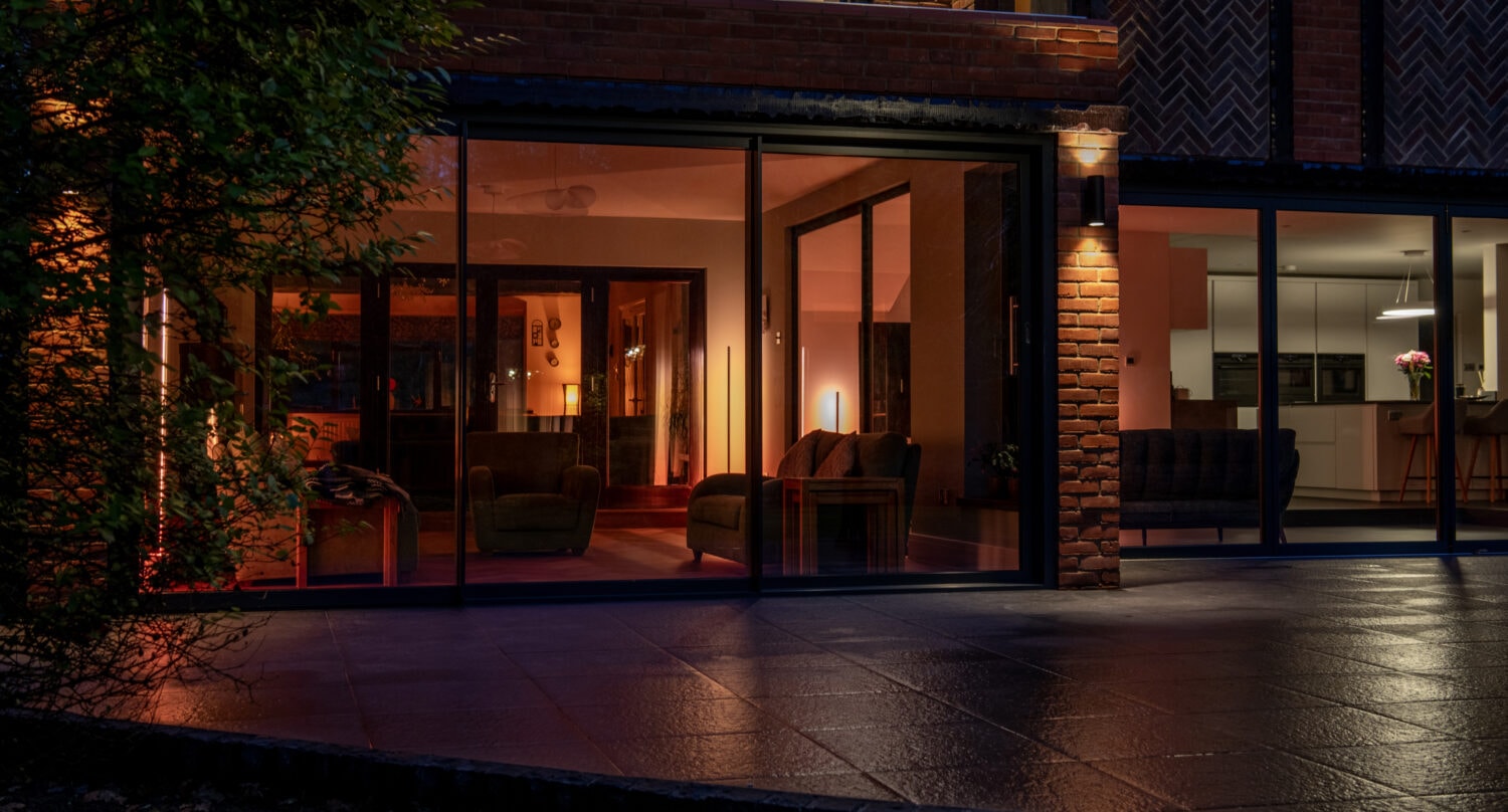 A modern house at night with large glass windows and sleek sliding doors revealing warmly-lit living and dining areas inside, featuring armchairs, sofas, and visible indoor plants. The exterior patio is dimly illuminated.