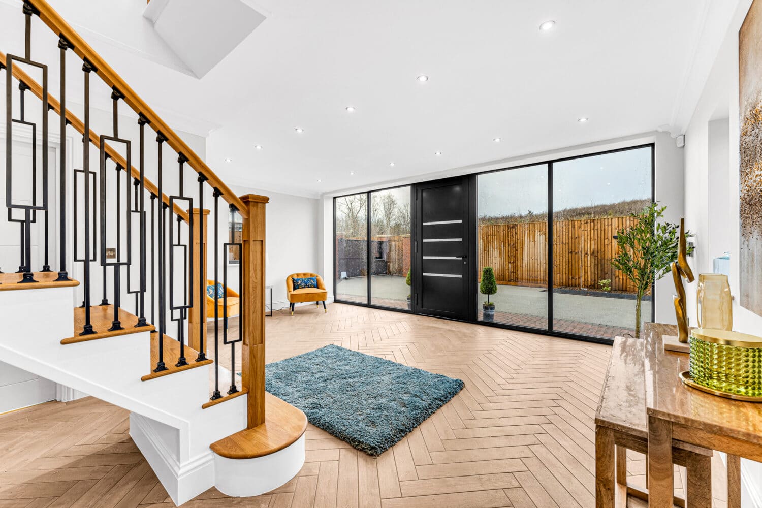 Modern, bright entryway with large black-framed front doors, a wooden staircase with black railings, a blue rug, a chair, and a wooden console table, featuring herringbone wood floors and an outdoor view.
