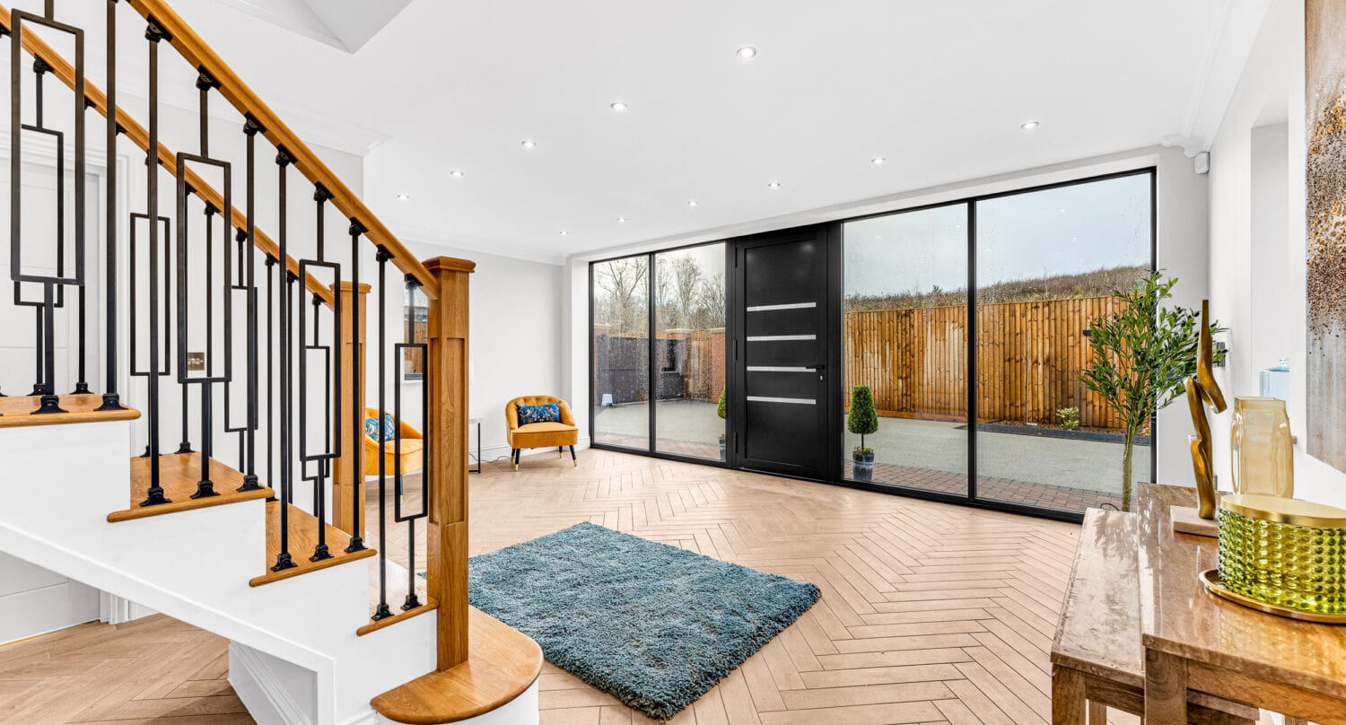 Modern, bright entryway with large black-framed front doors, a wooden staircase with black railings, a blue rug, a chair, and a wooden console table, featuring herringbone wood floors and an outdoor view.