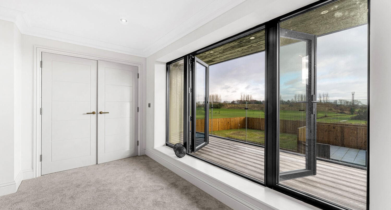 A bright, modern room with light carpet, white double doors, and large black-framed sliding doors opens to a wooden deck. The windows overlook a fenced green field and trees under a cloudy sky.