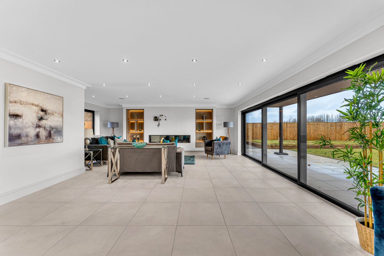 Spacious modern living room with large tiled floor, bespoke glass solutions like bifold doors opening to a patio, neutral walls, contemporary furniture, built-in shelves, and indoor plants. Natural light fills the room.