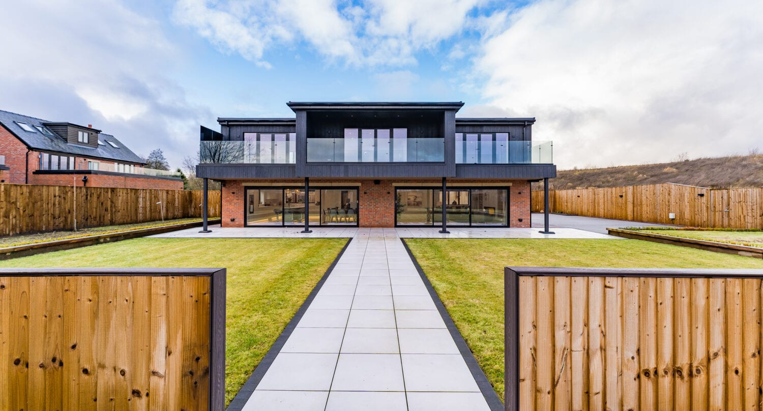 A modern two-story house with large glass windows, bespoke glass solutions, and a balcony, viewed from an open wooden gate with a paved path leading through a green lawn. The house is surrounded by a wooden fence and partly cloudy sky.
