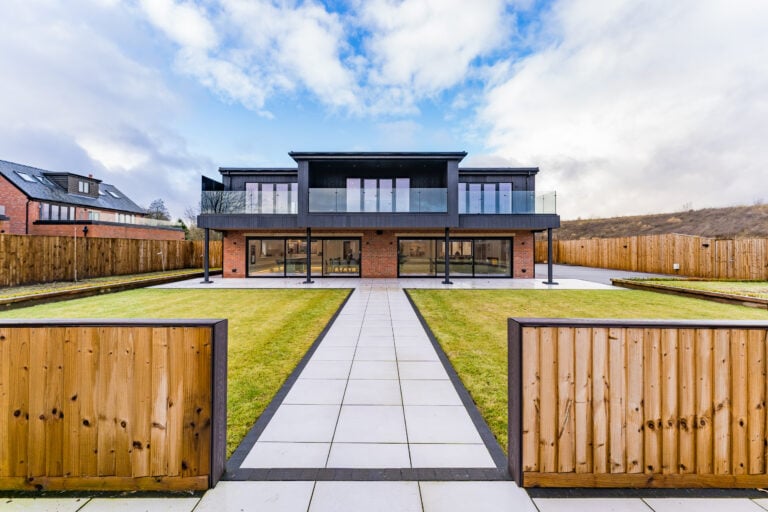 A modern two-story house with large glass windows, bespoke glass solutions, and a balcony, viewed from an open wooden gate with a paved path leading through a green lawn. The house is surrounded by a wooden fence and partly cloudy sky.