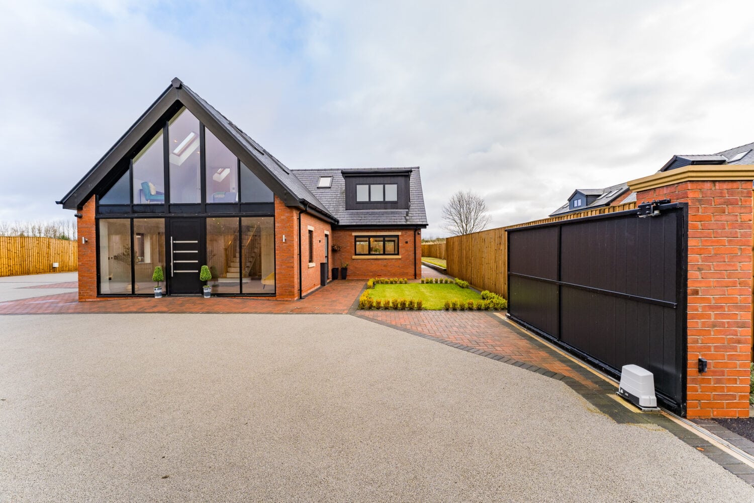 A modern brick house with large floor-to-ceiling windows and a sleek glass roofing, a sloped roof, paved driveway, and black sliding gate. Small shrubs line the grass yard along a wooden fence beneath a partly cloudy sky.