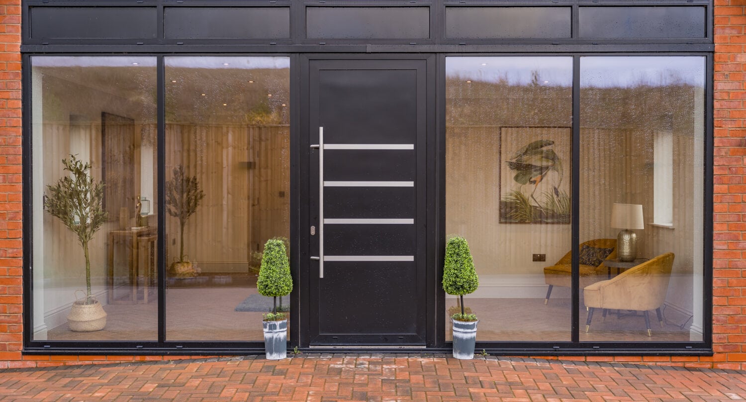 Modern house entrance with large glass windows, sleek bifold doors featuring horizontal metal accents, and two potted topiary plants on either side. Inside, a chair, lamp, and wall art are visible. Brick pavement in front.