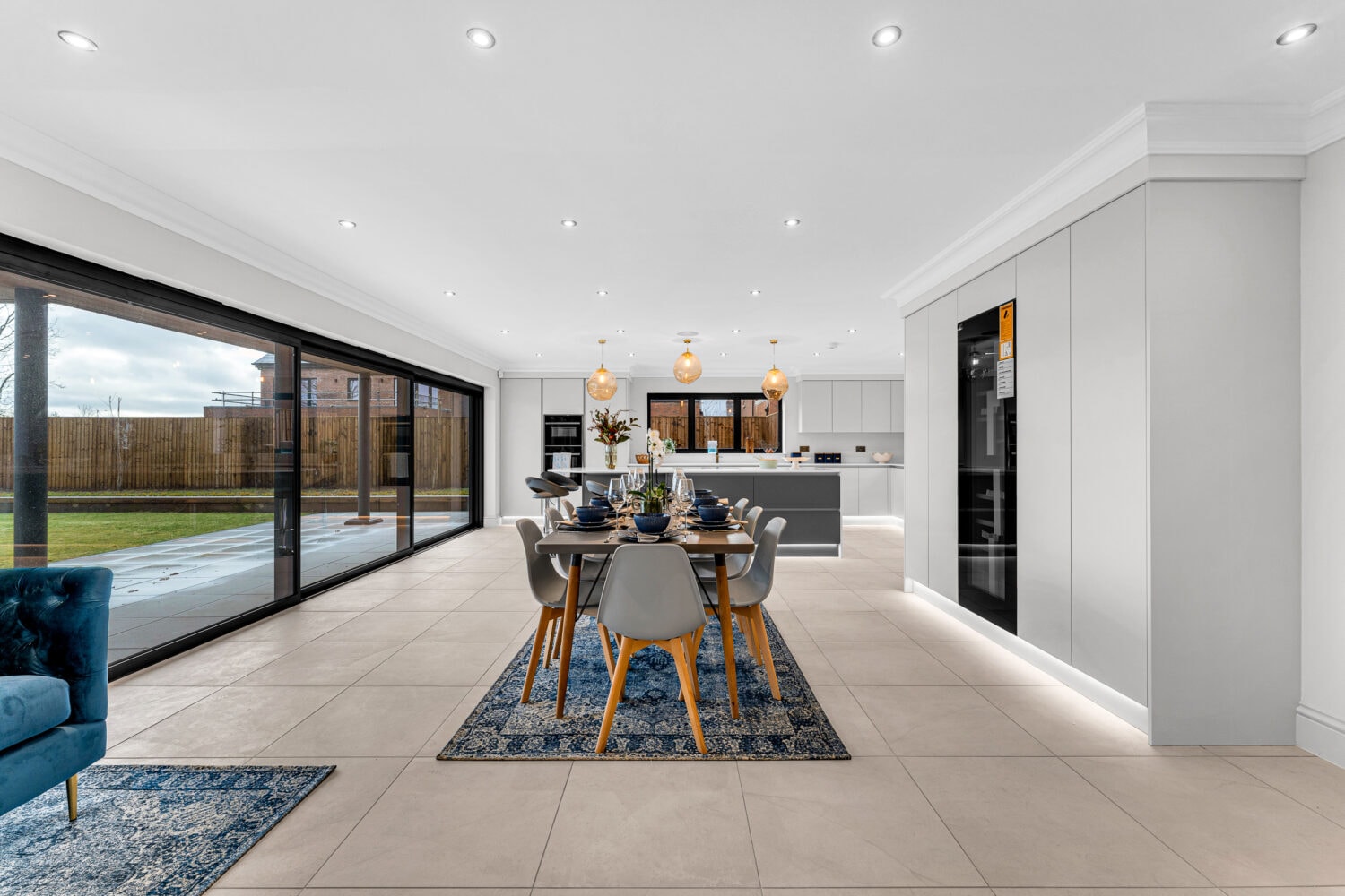 Modern open-plan kitchen and dining area with bespoke glass solutions, featuring floor-to-ceiling sliding doors, a table for six, white kitchen with pendant lights, bright tiled floors, and minimalistic decor.