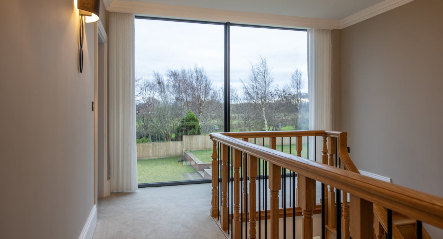 A hallway with beige walls and carpet, featuring a wooden railing and a large floor-to-ceiling window overlooking a green backyard with trees, bespoke glass solutions, and a wooden fence.