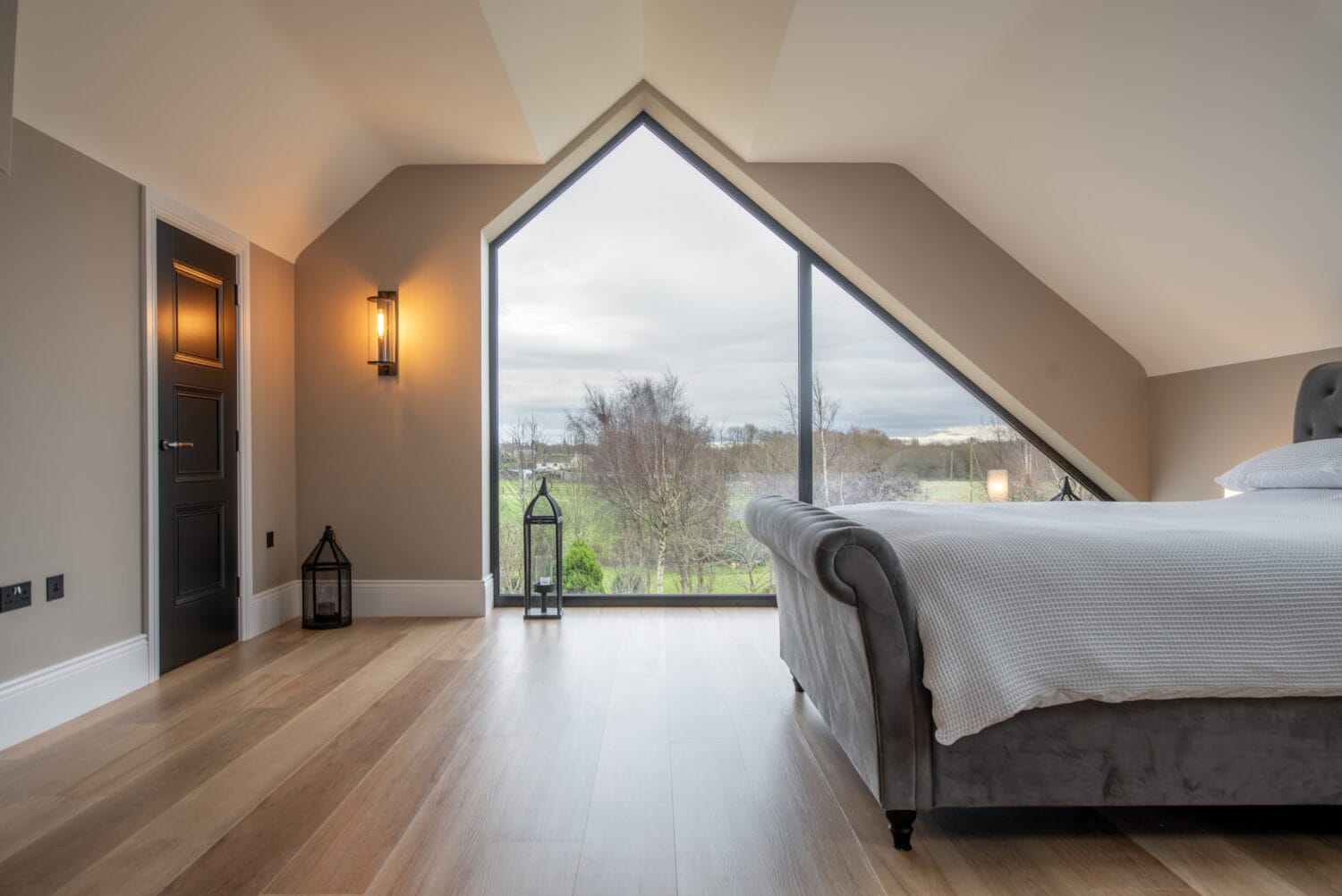Modern bedroom with sloped ceiling, large triangular window featuring bespoke glass solutions overlooking greenery, wooden floor, a dark bed with white bedding, wall lamp, and two lanterns by the window.