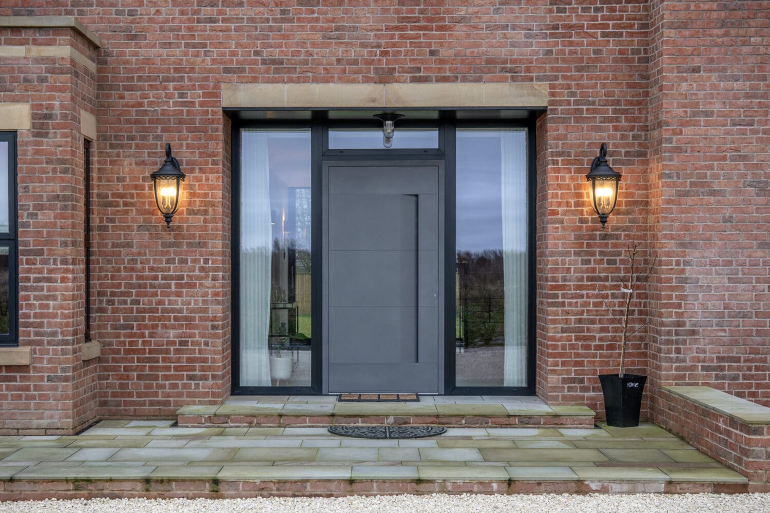 Modern gray front door with large glass side panels, set in a red brick wall. Two lit black lanterns flank the entrance. Stone steps and a doormat lead up to these stylish front doors.