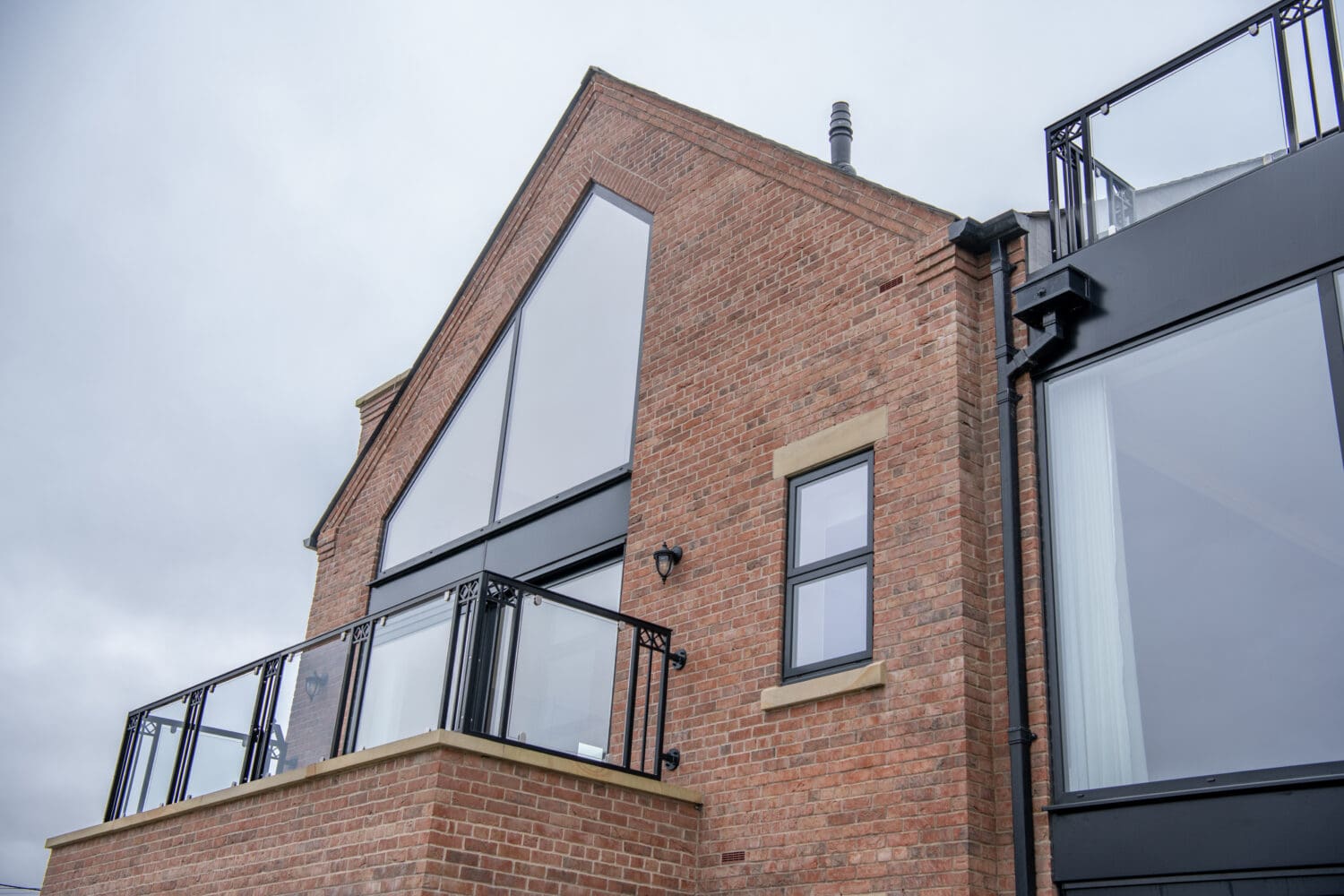 Modern brick house with large triangular and rectangular windows, black frames, glass balcony railing, and sleek sliding doors, set against a cloudy sky.