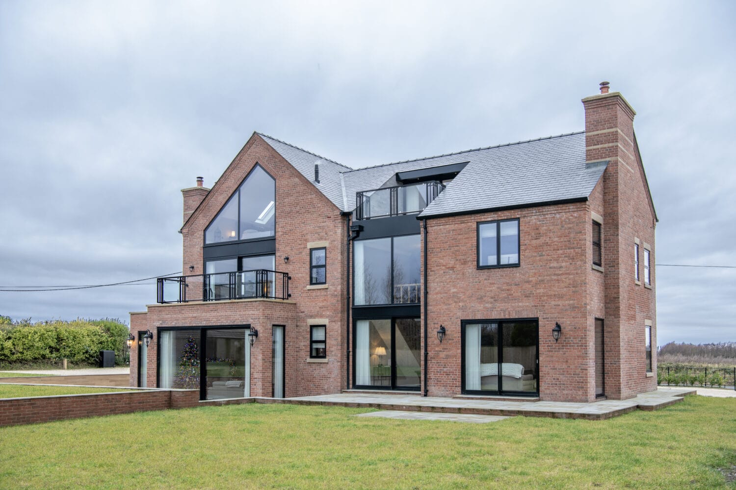 A modern three-story brick house with large glass windows and sliding doors, a rooftop balcony, and a sloped gray roof, set in a grassy yard with overcast skies in the background.