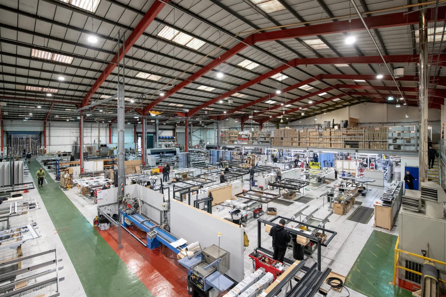 A large, modern factory interior with high ceilings, red beams, and bright lighting. Multiple workstations are arranged in rows, as workers assemble materials. The space features glass roofing and shelves and equipment line the walls.