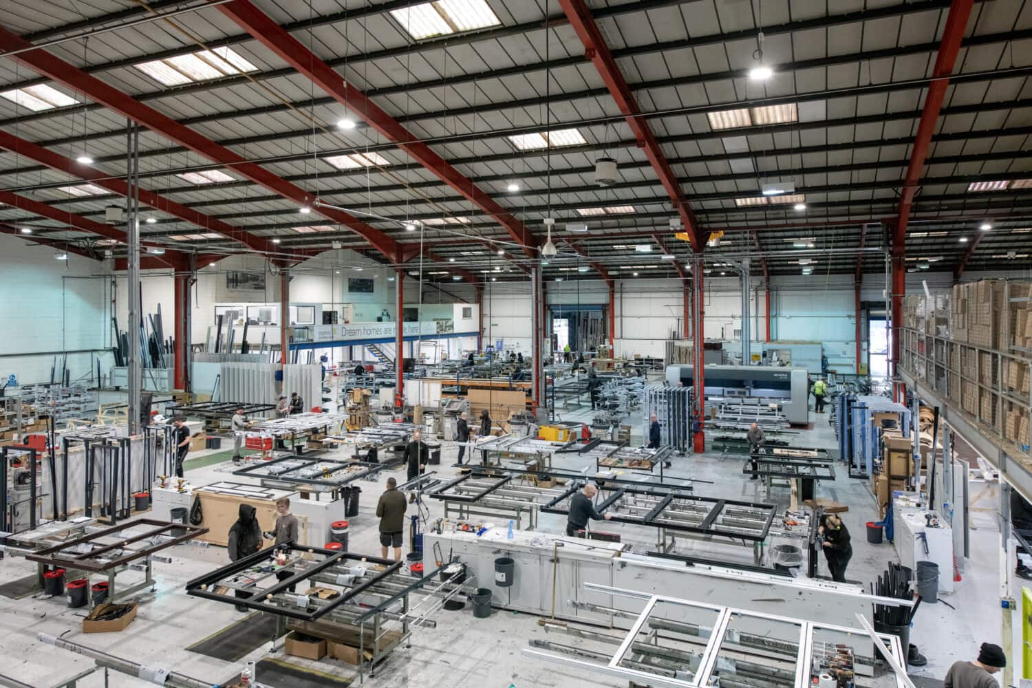 A spacious factory interior with high ceilings, red beams, and workers assembling large metal or glass frames for sliding doors at various workstations, surrounded by machinery and materials.