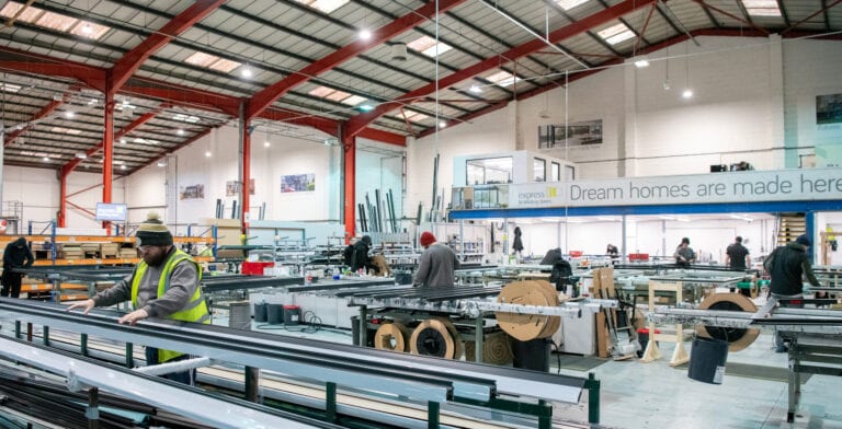 A spacious factory with workers assembling metal frames, including sliding doors, surrounded by machinery and large spools. The interior has high red steel beams, and a banner reads, Dream homes are made here.