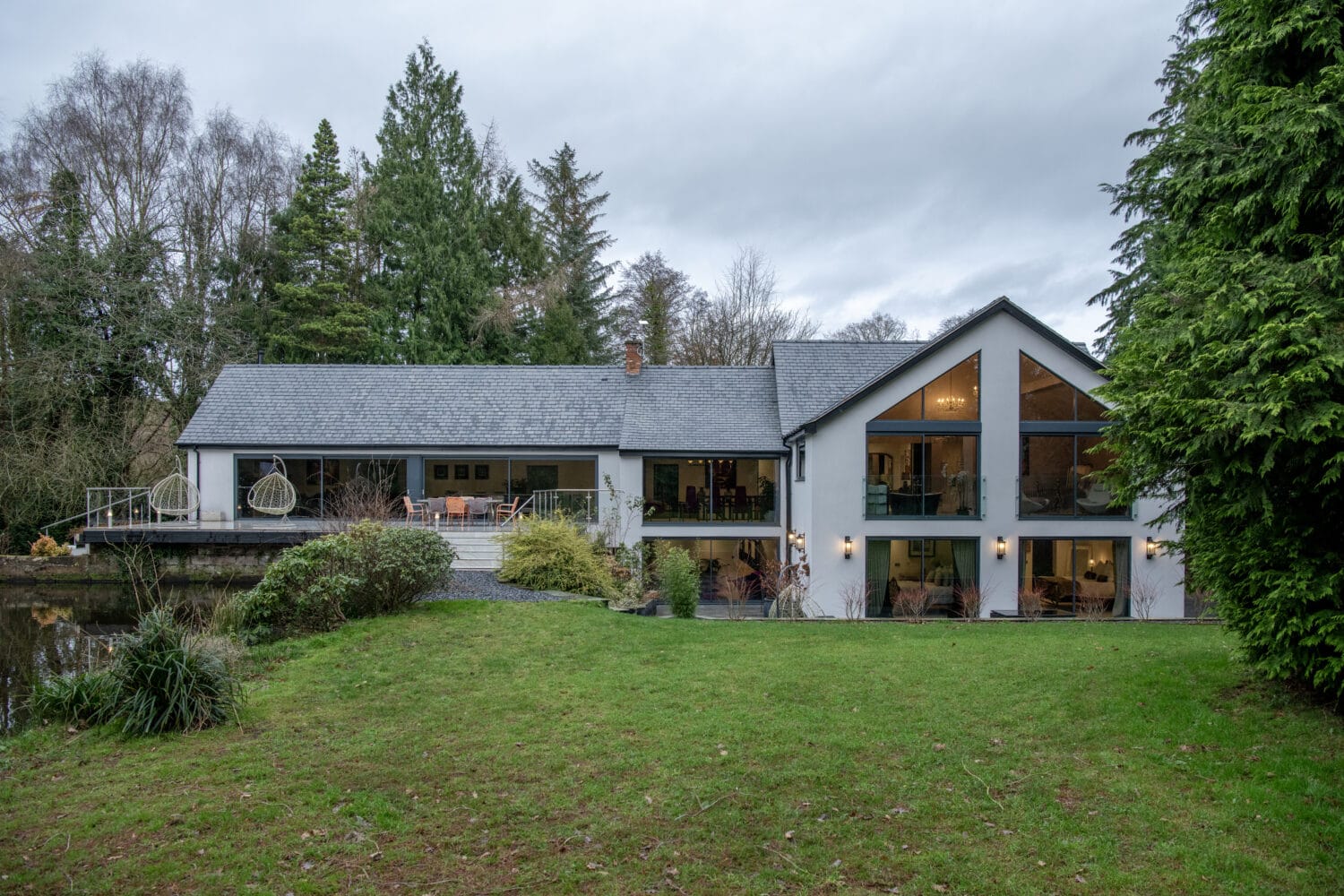 Modern two-story house with large glass windows and stylish bifold doors, surrounded by trees and greenery. The house features a spacious deck, outdoor seating, and sits on a well-kept lawn under an overcast sky.