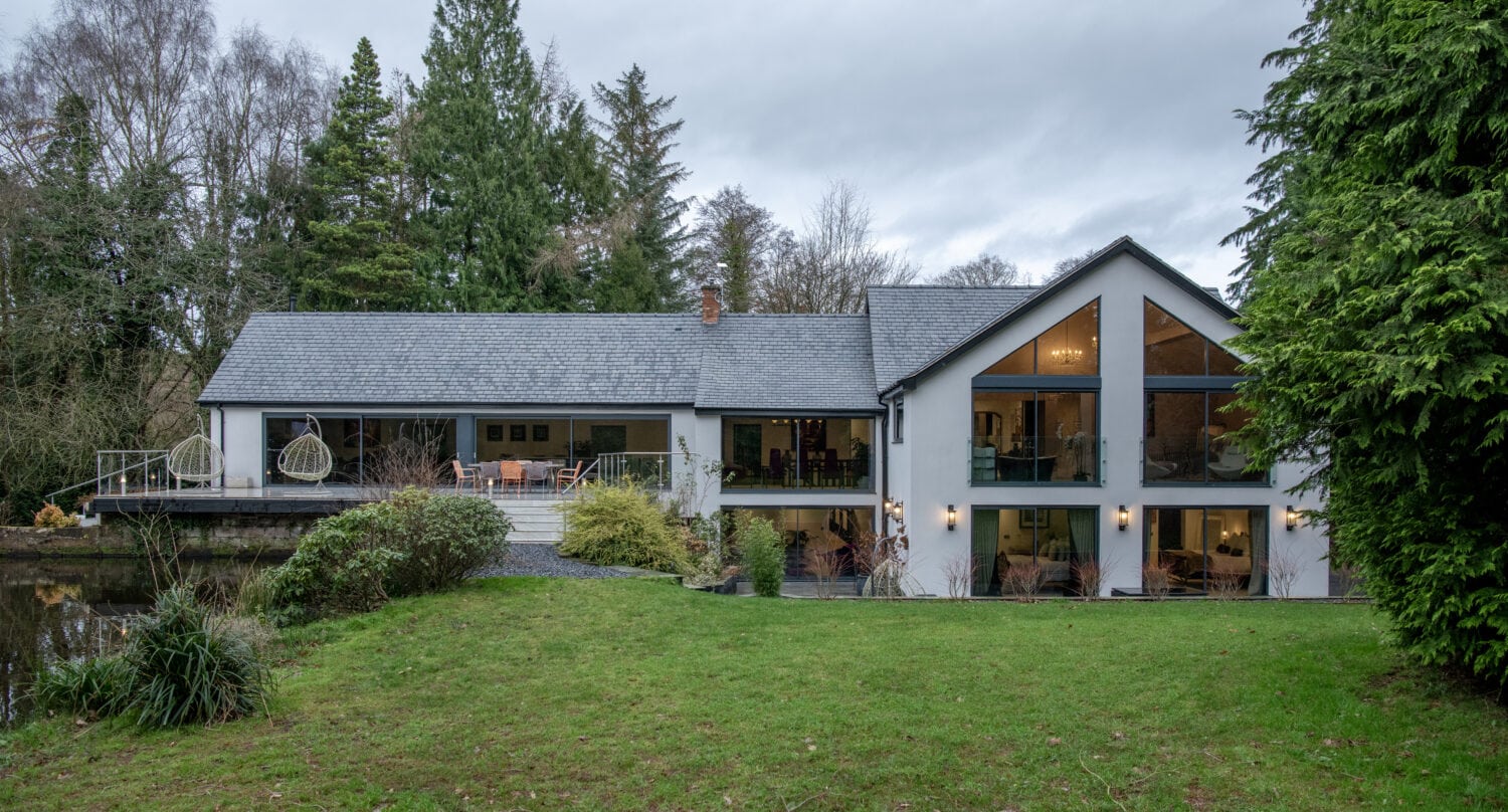 Modern two-story house with large glass windows and stylish bifold doors, surrounded by trees and greenery. The house features a spacious deck, outdoor seating, and sits on a well-kept lawn under an overcast sky.