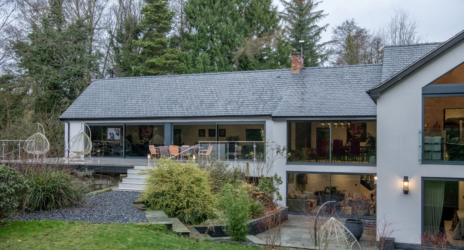 Modern two-story house with bespoke glass solutions, featuring large windows and bifold doors. Surrounded by lush greenery, it boasts an elevated patio with outdoor seating, a landscaped garden, and a grassy lawn in the foreground.