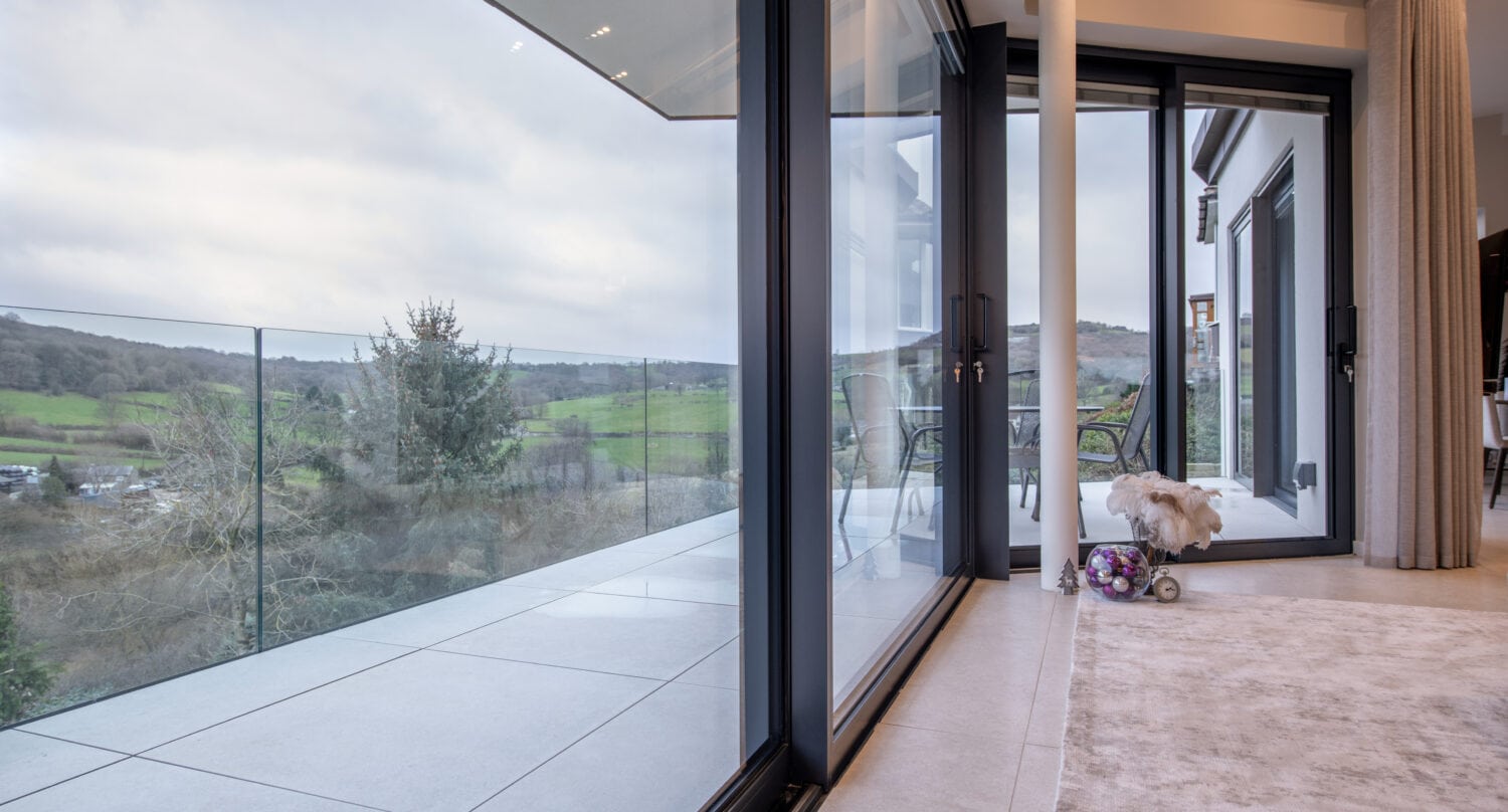 Floor-to-ceiling bespoke glass solutions sliding doors open to a balcony with glass railings, overlooking green hills and trees. Inside, a small dog sits on the carpet near a plush toy, close to the patio.
