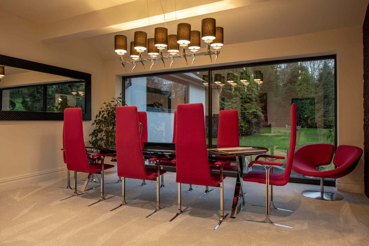 A modern dining room with a glass table, eight tall red chairs, a large window overlooking a green garden, and bespoke glass solutions like a rectangular mirror on the wall. A chandelier hangs above the table, enhancing the elegant space.