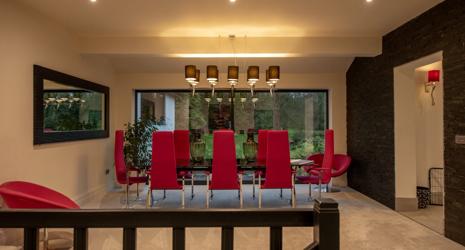 Modern dining room with a glass table, eight tall red chairs, matching armchairs, a large window with garden view, pendant lights, and a large mirror on the beige wall—enhanced by bespoke glass solutions for a sleek finish.