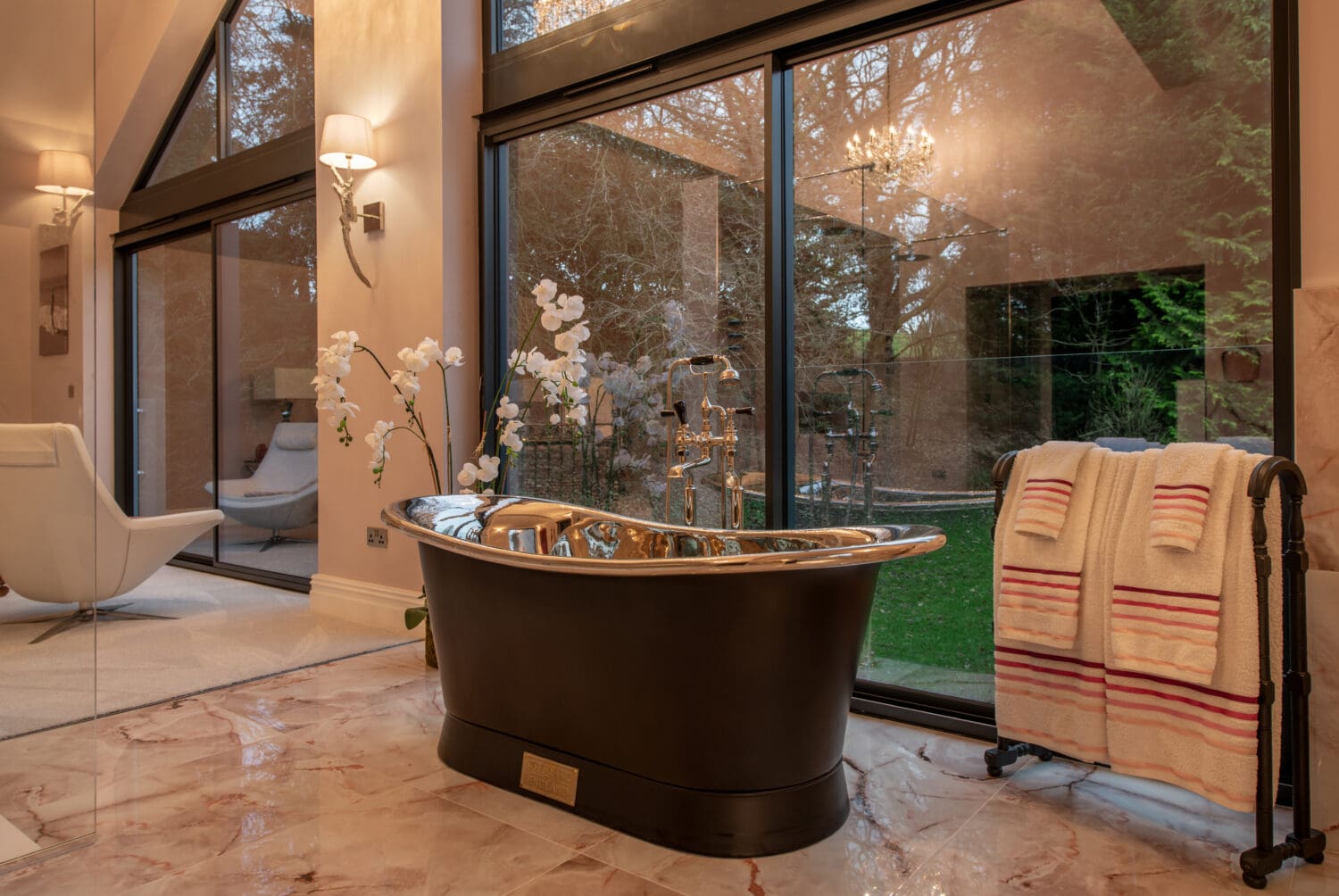 A luxurious bathroom with a freestanding black and silver bathtub, marble flooring, and a towel rack holding striped towels. Floor-to-ceiling windows and bespoke glass solutions reveal a green garden, while an orchid plant adds elegance to the space.