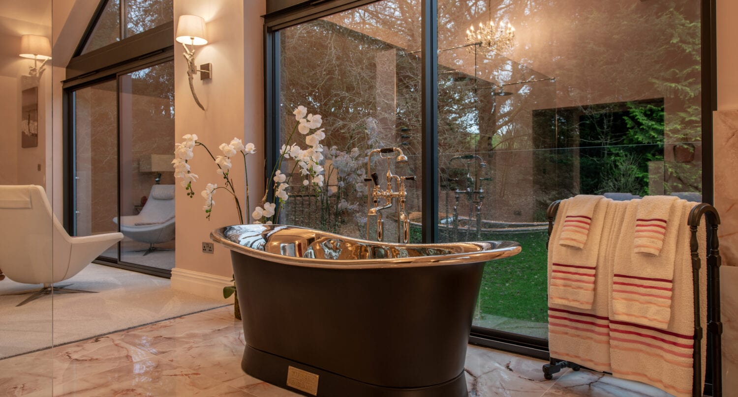 A luxurious bathroom with a freestanding black and silver bathtub, marble flooring, and a towel rack holding striped towels. Floor-to-ceiling windows and bespoke glass solutions reveal a green garden, while an orchid plant adds elegance to the space.