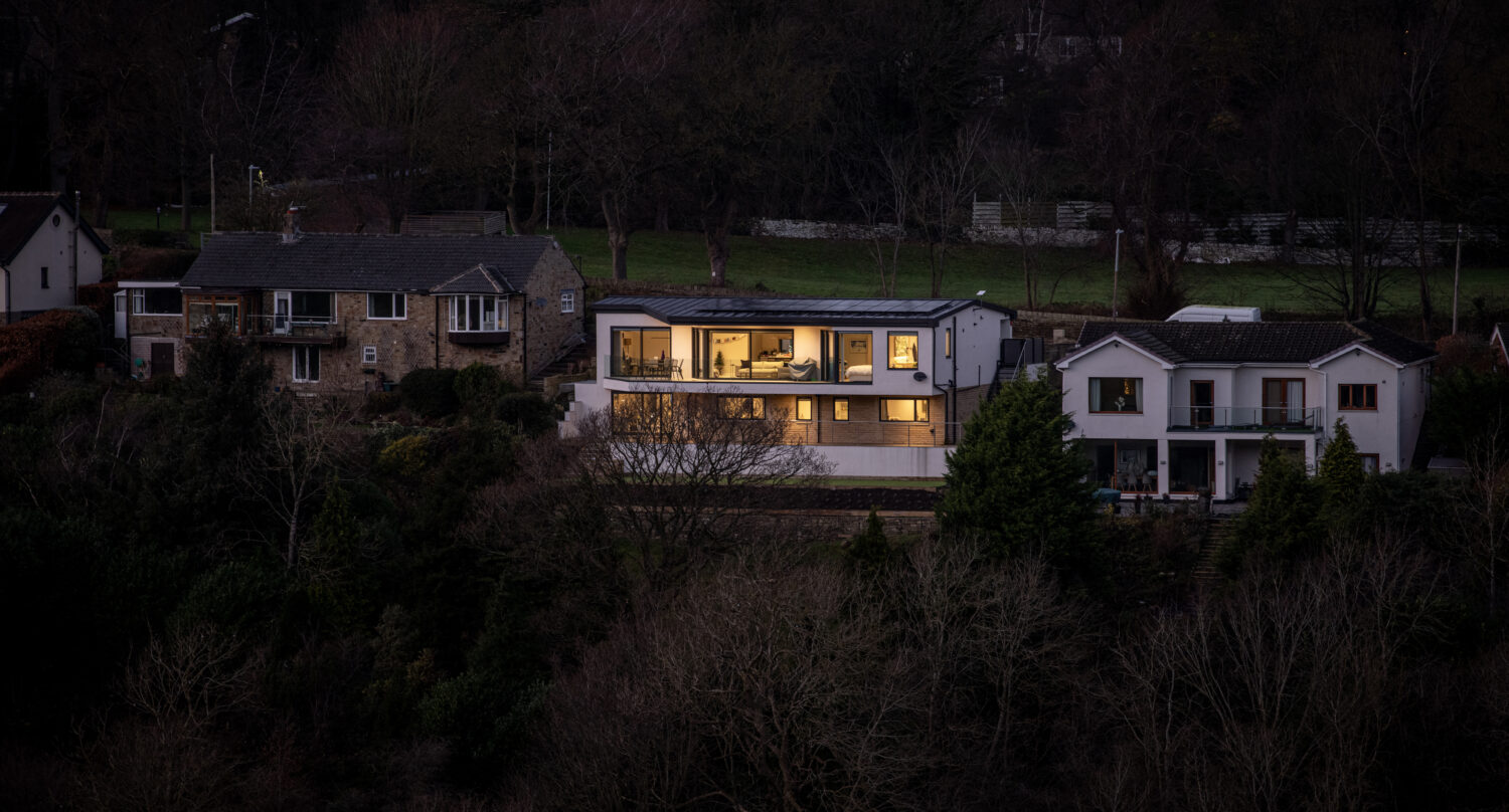 A modern house with brightly lit windows and sleek bifold doors stands between two traditional homes at dusk, surrounded by dense trees and dark woodland in the background.