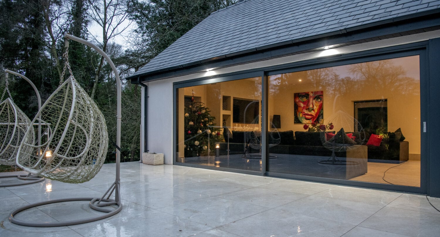 Modern house with large glass sliding doors and glass roofing opens onto a tiled patio with two hanging wicker chairs. Inside, a decorated Christmas tree and a colorful portrait brighten the cozy living room. Trees surround the house.
