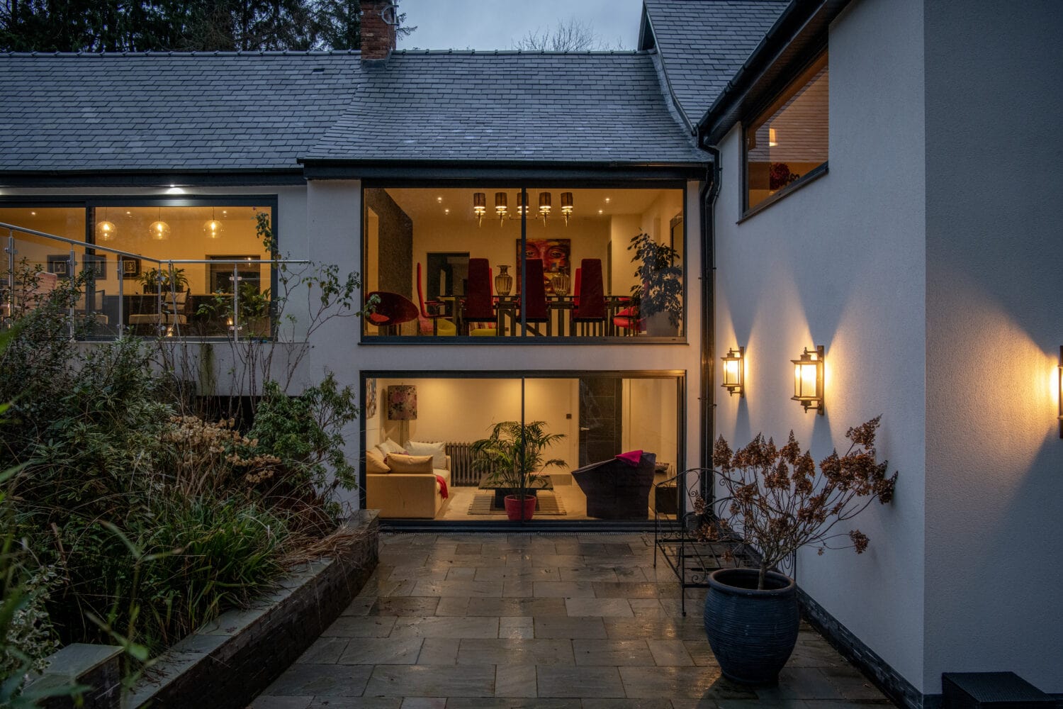 Modern house exterior at dusk with large windows, bespoke glass solutions front doors, warmly lit interior rooms, contemporary furniture, wall lights, potted plants, and a stone patio surrounded by greenery.