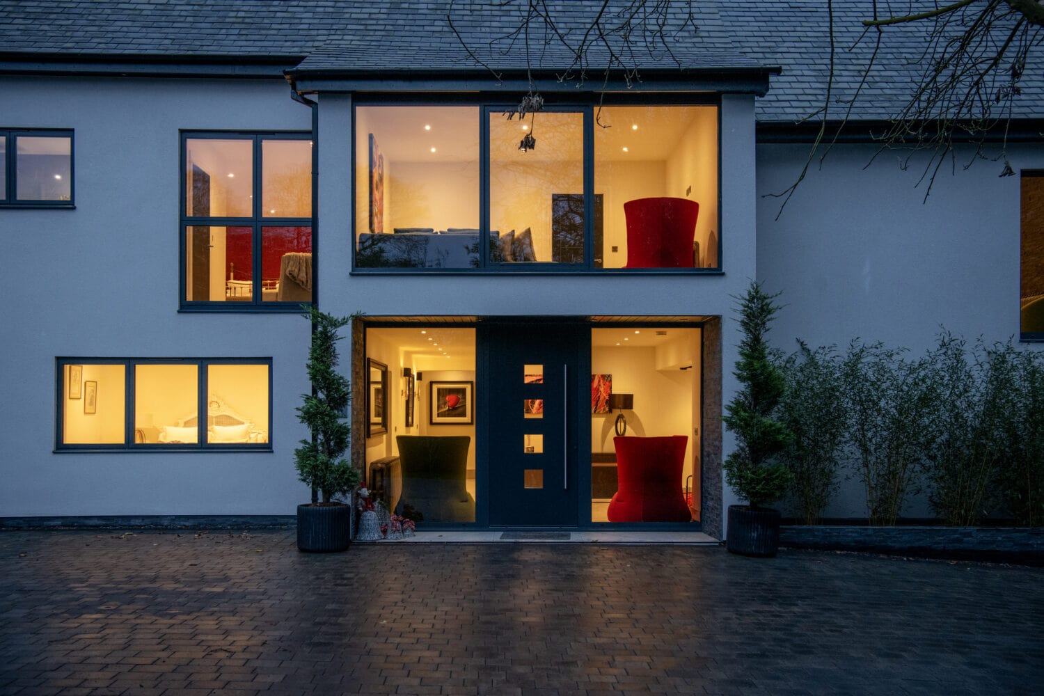Modern two-story house at dusk, with large windows glowing warmly from interior lights; minimalist design features sliding doors, light walls, potted plants, and a paved driveway.