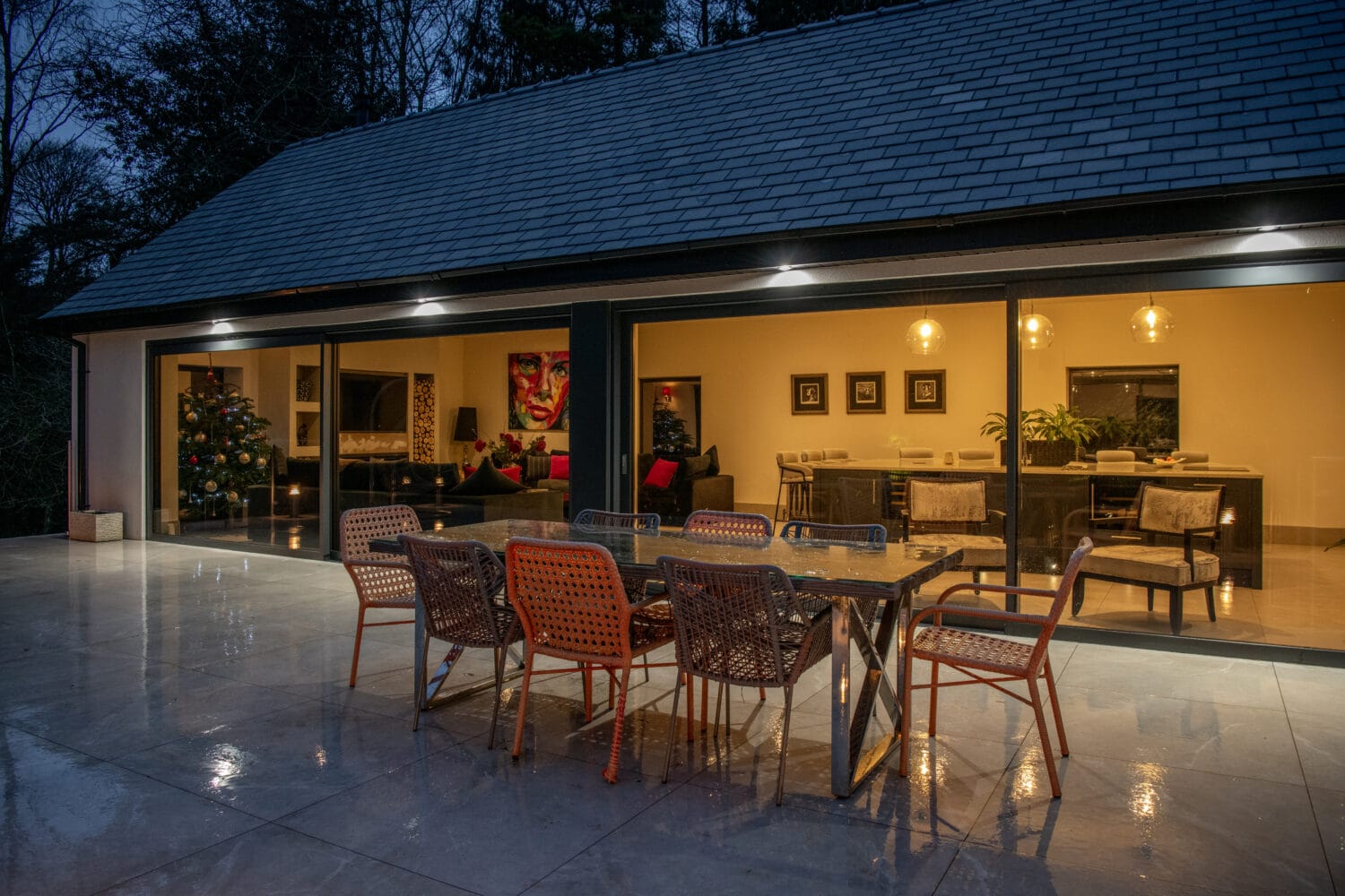 Modern patio at dusk with a glass dining table and colorful chairs on a wet tiled floor. Warm light fills the living area inside, where a Christmas tree and art are visible through bespoke glass solutions and large sliding doors.