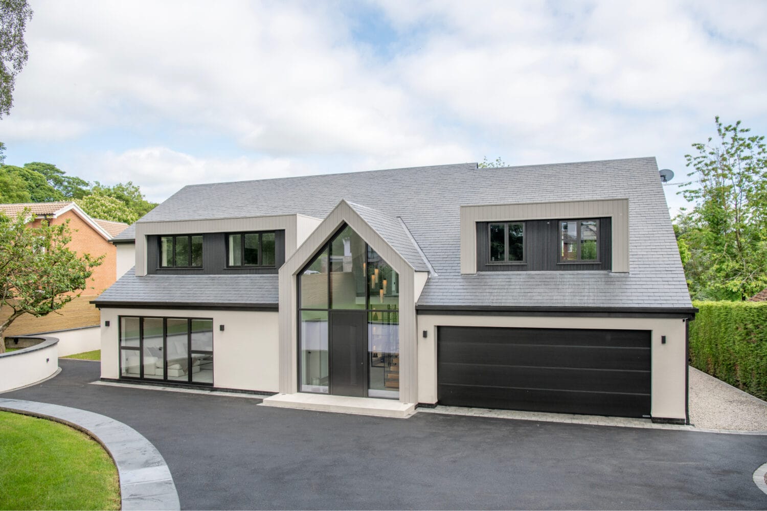 Modern two-story house with large windows, gray roof, and black double garage door. The exterior is cream and gray, featuring bespoke glass solutions and sleek sliding doors, with a driveway and well-kept lawn surrounding the property.