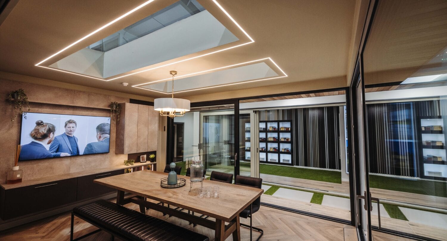 Modern meeting room with a wooden table, benches, and a TV displaying people in conversation. Large windows and sliding doors open to a green courtyard and shelves. Skylights and geometric lighting brighten the space.