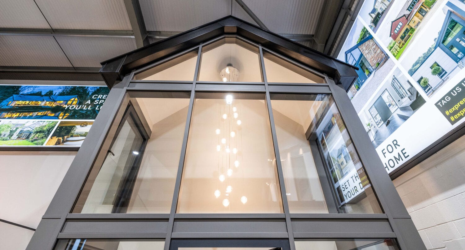 A modern glass entryway with a peaked glass roofing is shown from below, featuring a tall chandelier inside. Large posters with images of houses and text are displayed on the walls above.