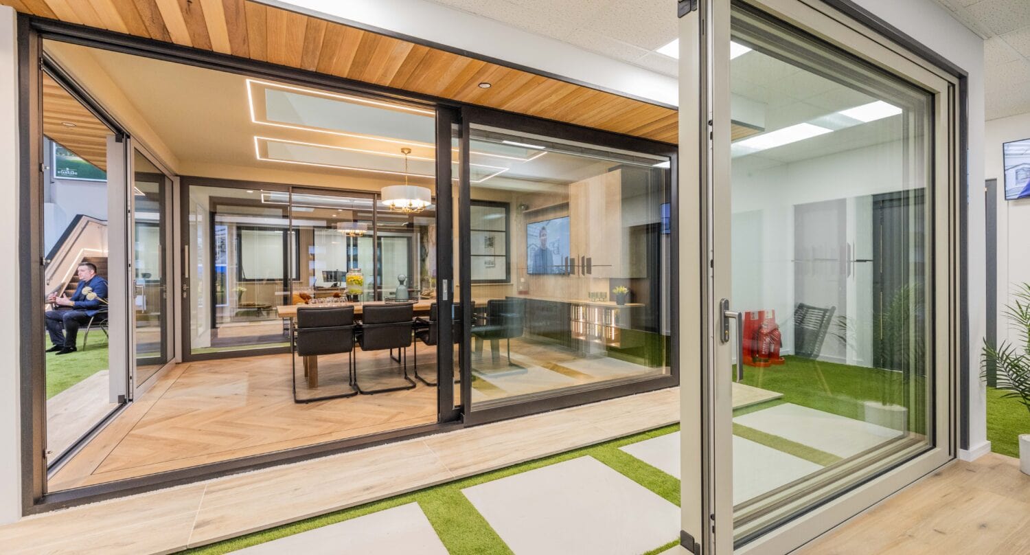 Modern office space featuring bespoke glass solutions with large glass walls and sliding doors, wood flooring, a conference table with chairs, and green artificial grass accents. A person sits in the background on the left side.