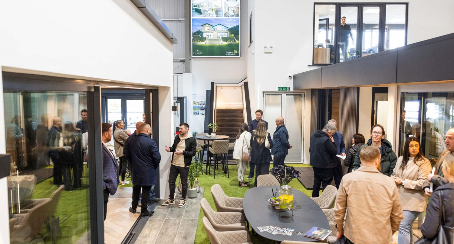 A group of people mingle and converse in a modern, open indoor showroom with large glass windows, bifold doors, tables with brochures, and bright lighting. Some are seated, while others stand, engaging in discussion.