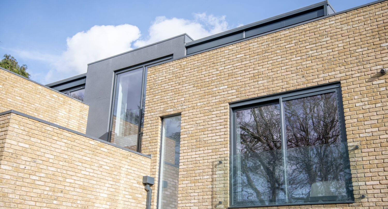 Modern building with light-colored brick walls, large rectangular windows, and bespoke glass solutions that reflect the blue sky with scattered clouds.