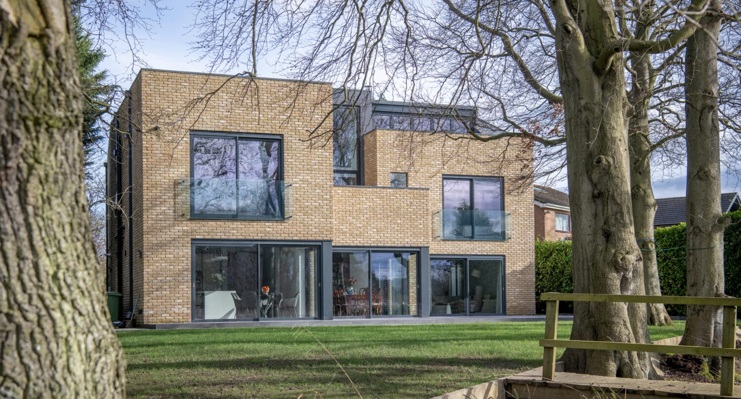 A modern two-story house with large glass windows, sliding doors, and a brick exterior, surrounded by grass, trees, and a small wooden deck in the foreground.