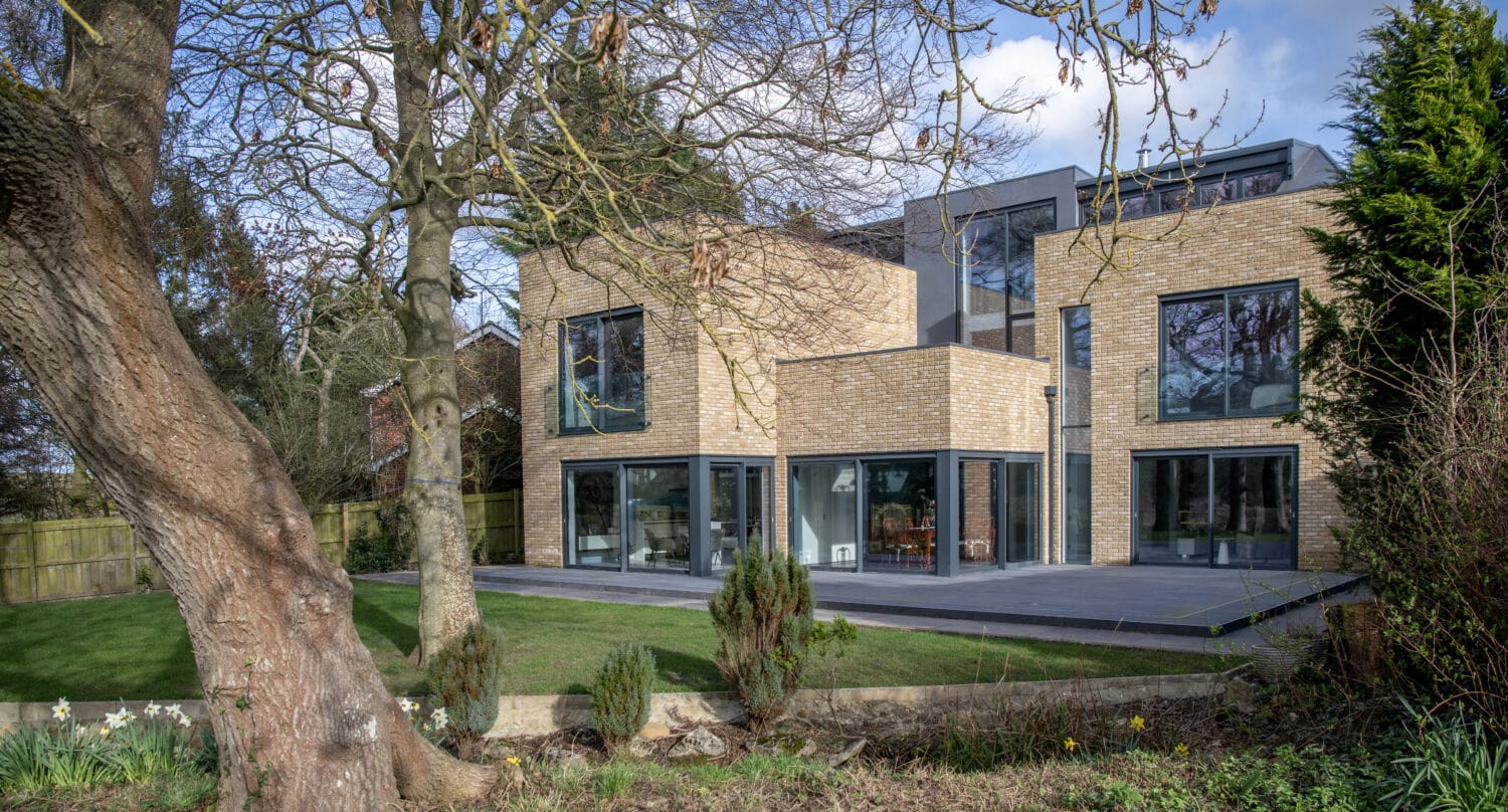 A modern two-story house with large glass windows, light brick walls, and sleek bifold doors, set in a green backyard with trees, shrubs, and a wooden fence on a partly cloudy day.