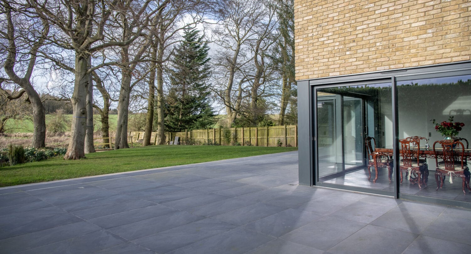 Modern house with bespoke glass solutions, featuring large sliding doors opening onto a spacious gray tiled patio that overlooks a grassy yard with leafless trees and a wooden fence. A dining table with chairs is visible inside.