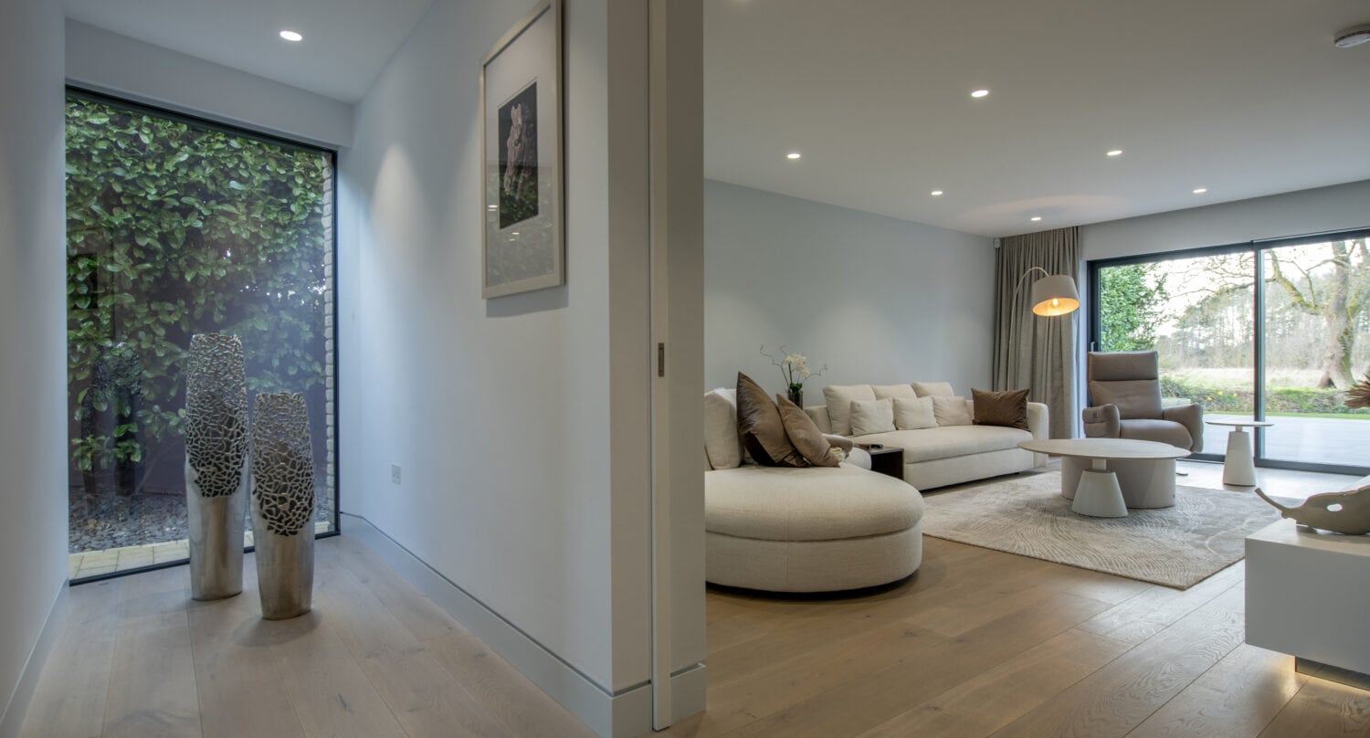 A modern living room with large windows, bespoke glass solutions, neutral-colored furniture, a round ottoman, and a hallway with wooden floors and decorative sculptures. Trees and greenery are visible outside.